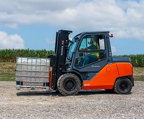 Forklift with orange and dark blue body lifting a white pallet crate in an outdoor industrial setting with cornfield in background