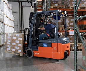 Operator in forklift lifting stacked cardboard boxes and pallets in warehouse with shelving and inventory in background