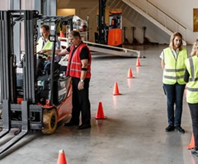 Instructor in yellow safety vest observing trainee operating red forklift in warehouse with orange traffic cones and safety equipment
